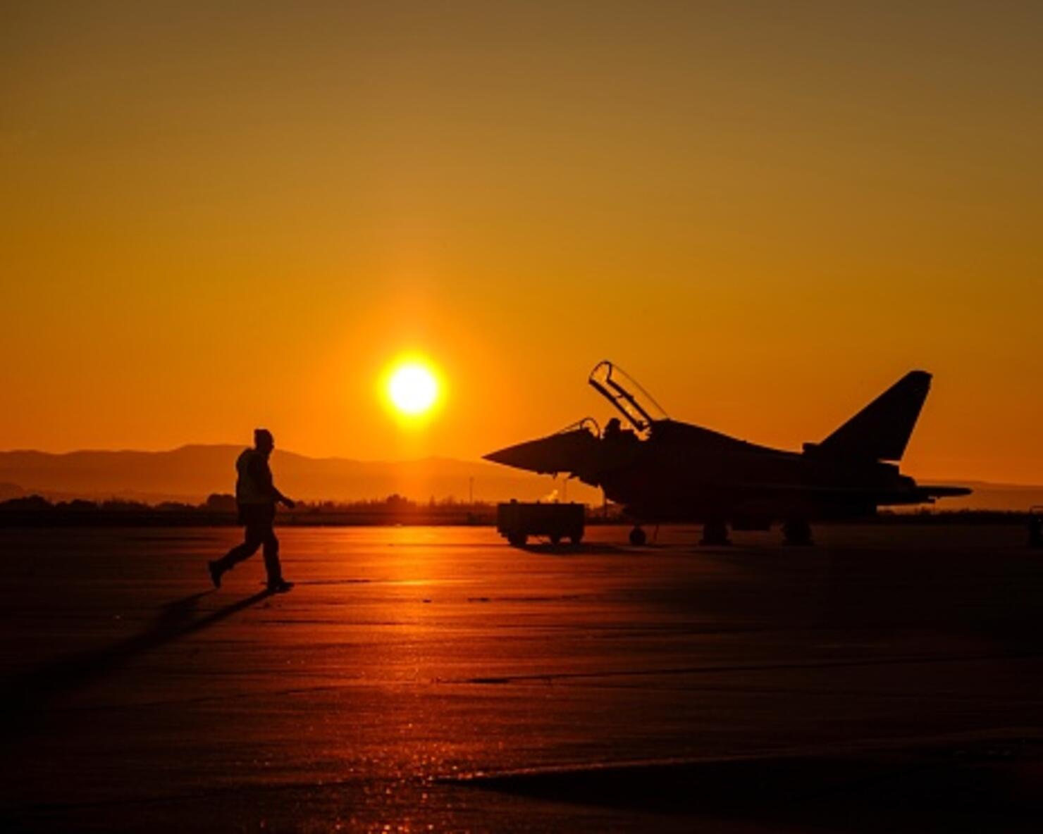 Silhouette Man Walking By Fighter Plane On Runway During Sunset