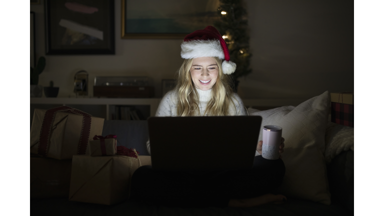 Young blonde woman in Santa hat using laptop in dark living room