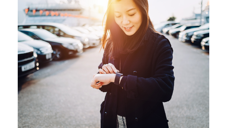 Beautiful young Asian woman checking time on smartwatch in city, in front of cars in outdoor carpark at sunset