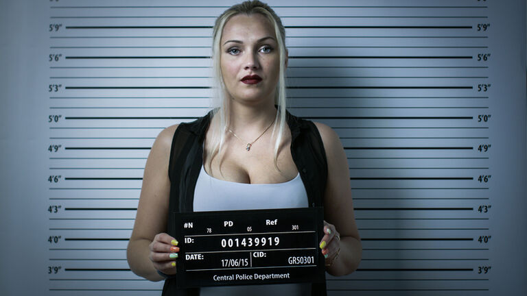 In a Police Station Arrested Woman Gets Front-View Mug Shot. She Wears Saucy Clothes, Has Heavy Makeup and Holds Placard. Height Chart in the Background. Shot with Dark Cold Lights and Vignette Filter