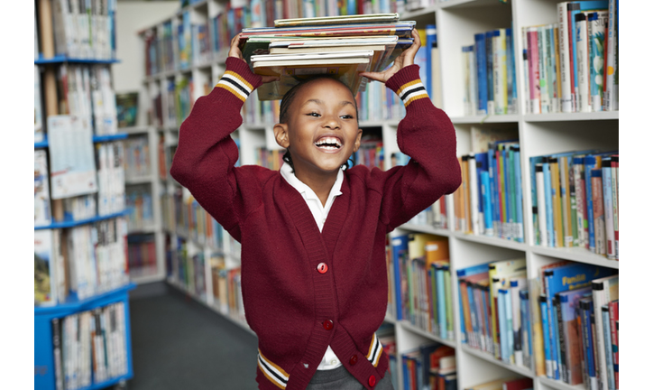 Cute schoolgirl smiling & balancing stack of books on the head at library