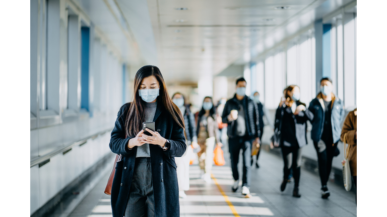 Asian woman with protective face mask using smartphone while commuting in the urban bridge in city against crowd of people