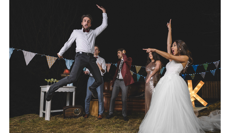 Surprised bride looking at man jumping on a night field party with friends