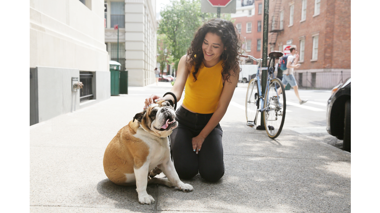 A LatinX female millennial sits next to a brown and white bulldog on the sidewalk of New York City.