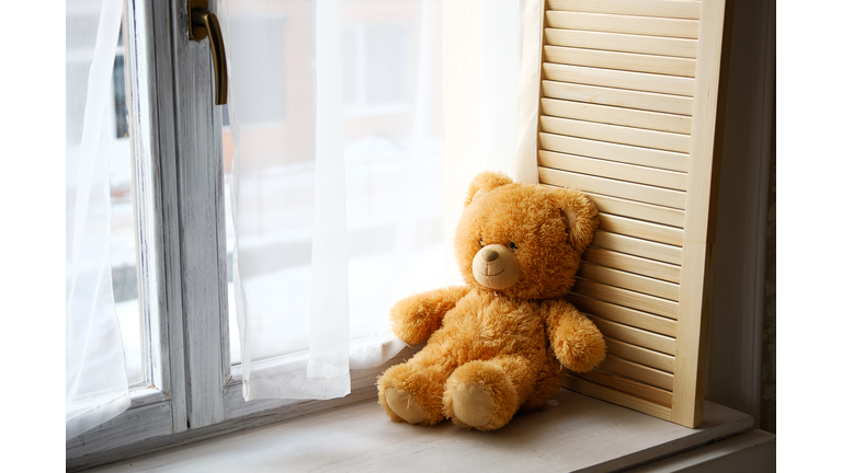 A cute little Teddy bear toy sits on a brown wooden and white plastic windowsill with a copy of the text space on the window glass. Beautiful lonely bear on the window. The concept of childhood.