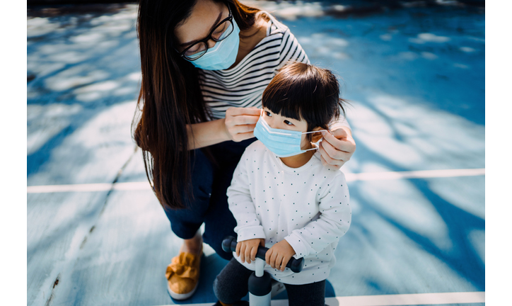 Young mother putting on surgical mask for little daughter in the playground to prevent the spread of cold and flu and viruses