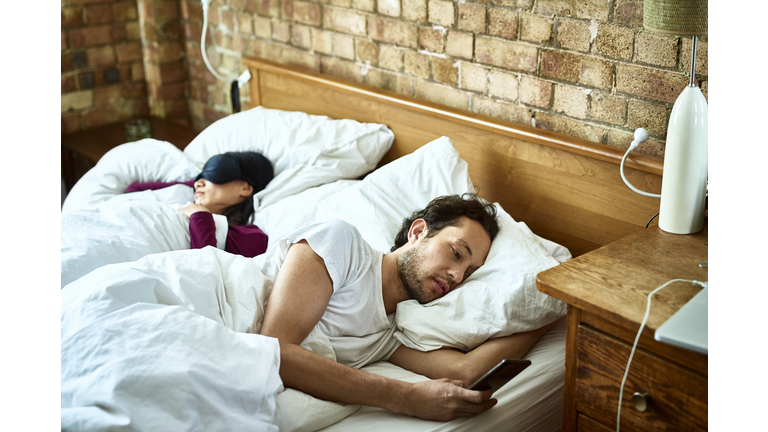 Woman fast asleep next to partner who is checking his smartphone