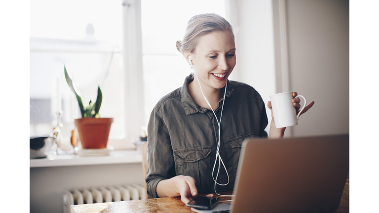 Happy woman listening to headphones while holding coffee cup at home