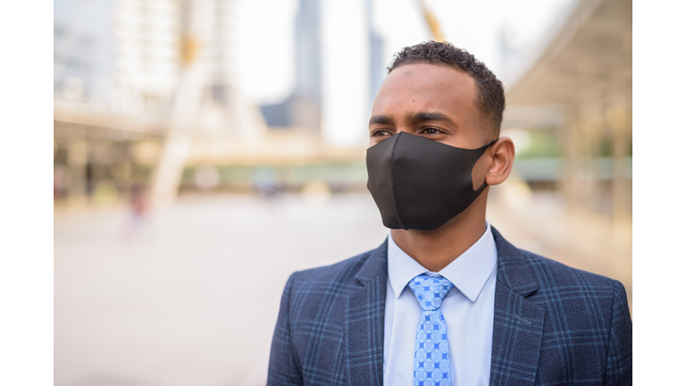 Face of young African businessman with mask for protection from corona virus outbreak thinking in the city