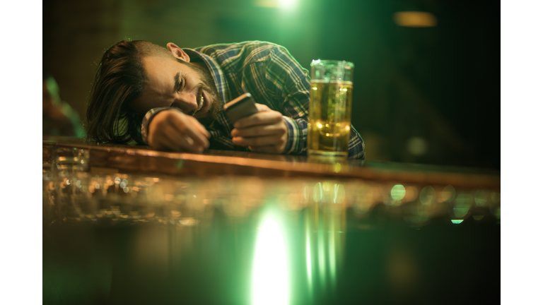 Young wasted man text messaging on cell phone while leaning on a bar counter.