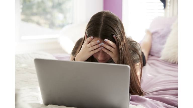 Upset teen girl lying on bed with head in hands using laptop