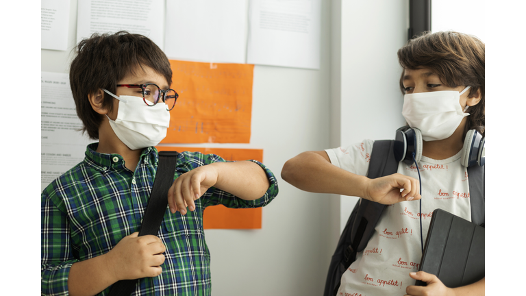 Boys wearing masks giving elbow bump while standing against wall in school