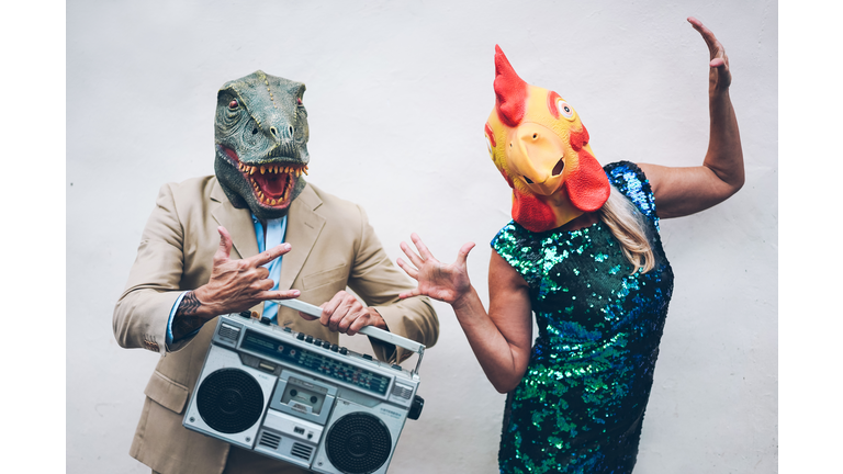 Crazy senior couple dancing for new year's eve party wearing t-rex and chicken mask - Old trendy people having fun listening music with boombox stereo - Absurd and funny trend concept - Focus on faces
