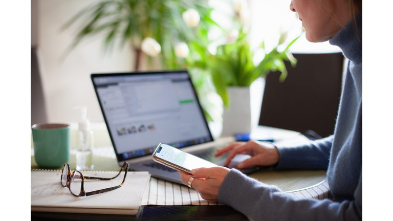 Woman working from home using laptop computer while reading text message on mobile phone