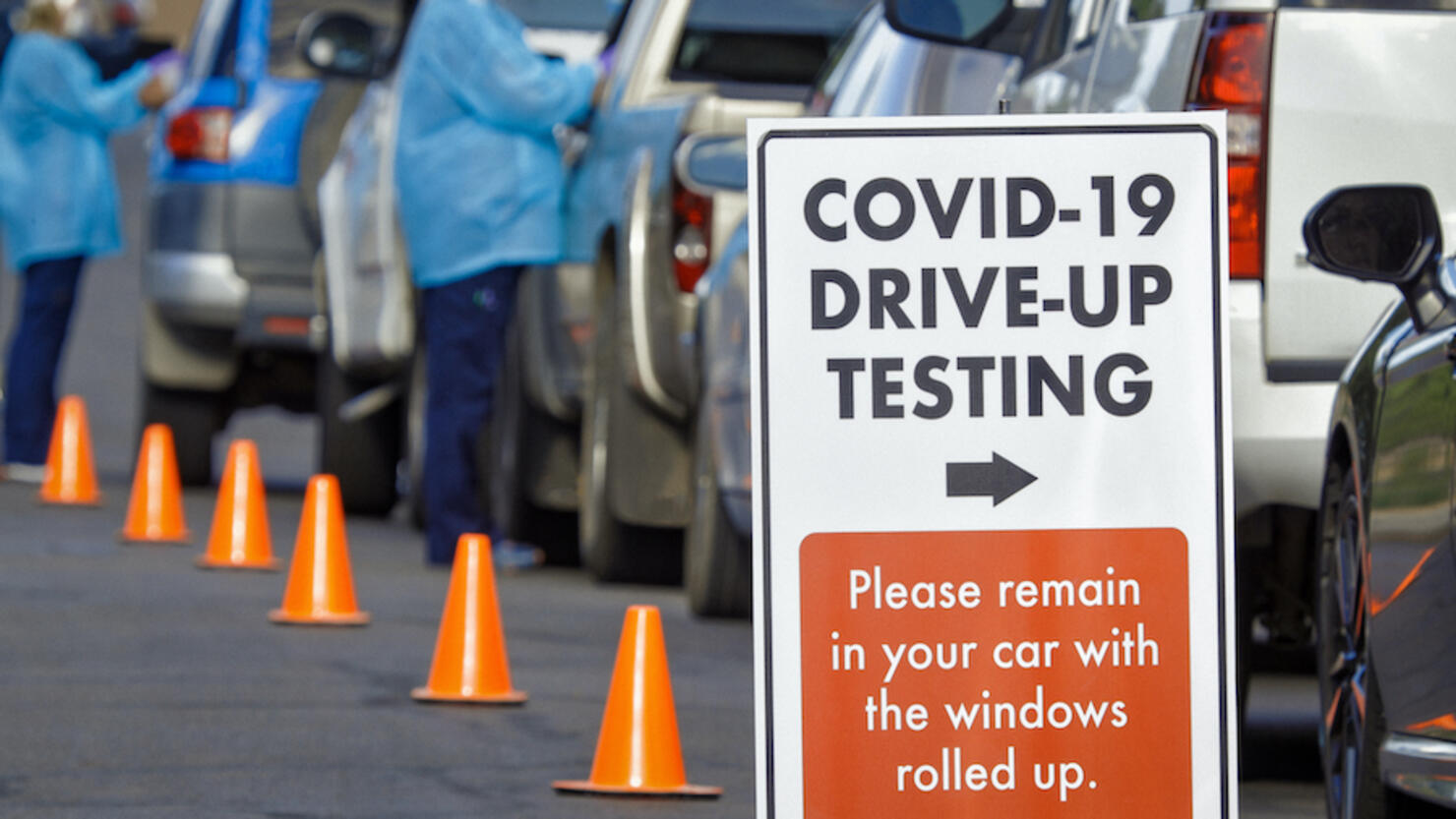 A "COVID-19 Drive-Up Testing" Sign Sits in the Foreground While Two Female Nurses Wearing Gowns and Surgical Face Masks Talk to Patients in their Cars in a Drive-Up (Drive Through) COVID-19 (Coronavirus) Testing Line Outside a Medical Clinic/Hospital Outd