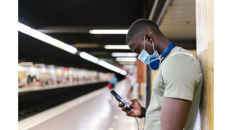 Young man wearing mask using smart phone while standing by wall in subway station