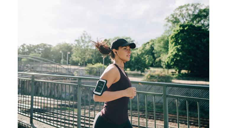 Confident sportswoman listening music through in-ear headphones while jogging on bridge in city