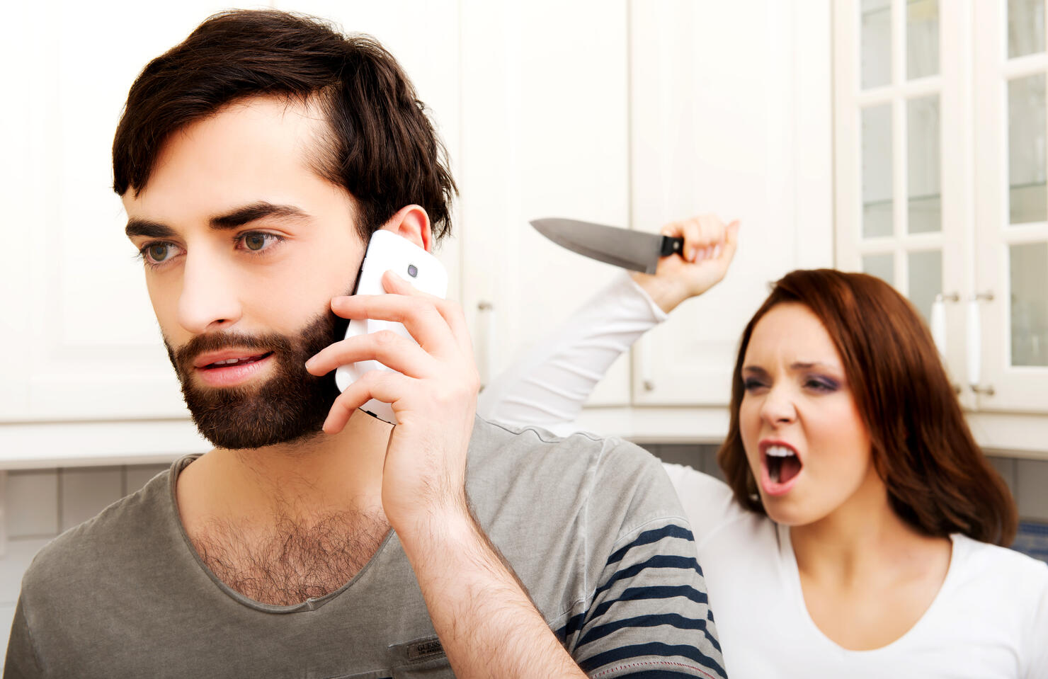 Angry Girlfriend Holding Knife Behind Boyfriend Talking On Phone At Home
