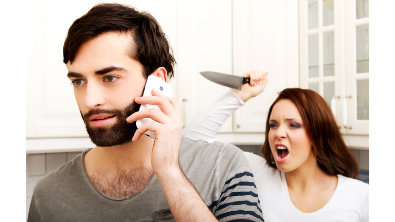 Angry Girlfriend Holding Knife Behind Boyfriend Talking On Phone At Home