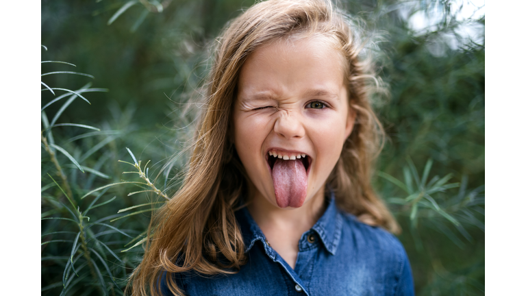 Front view portrait of small girl standing outdoors, sticking out tongue.