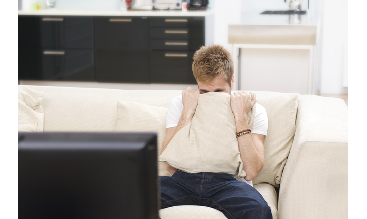 tense man watches television hiding behind cushion