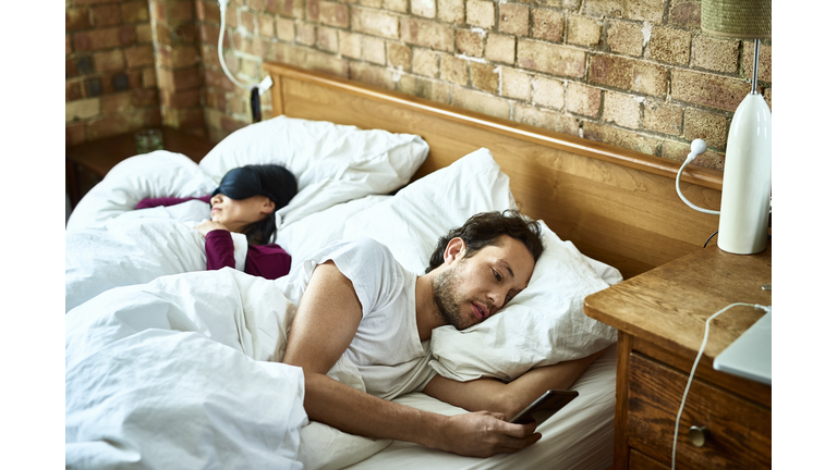 Woman fast asleep next to partner who is checking his smartphone