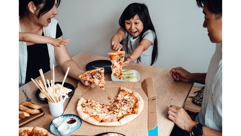 Cheerful Young Girl Holding A Slice Of Pizza, Sharing Meal With Family