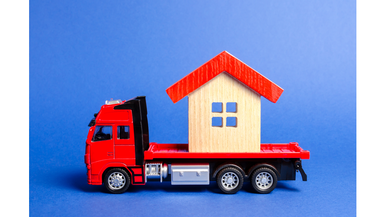 Red truck transports a red roofed house. Concept of transportation and cargo shipping, moving company. Construction of new houses and objects. Industry. Logistics and supply. Move entire buildings.