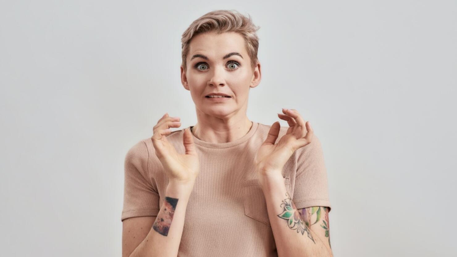 Portrait of tattooed woman with pierced nose and short hair in beige t shirt frowning face, feeling awkward, silly isolated over light background