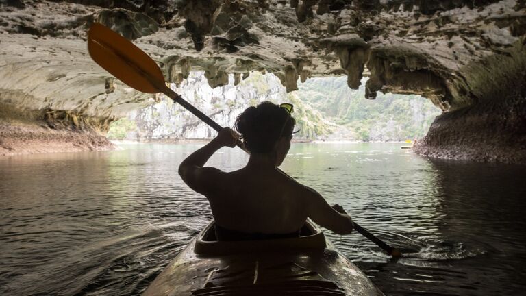 A rear view of a young woman paddling a yellow kayak through a cave in the Karst formations of Halong Bay.