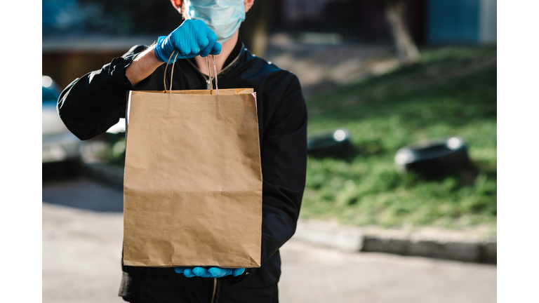 Courier in protective mask, medical gloves delivers takeaway food.  Employee hold cardboard package. Place for text. Delivery service under quarantine, 2019-ncov, pandemic coronavirus, covid-19.