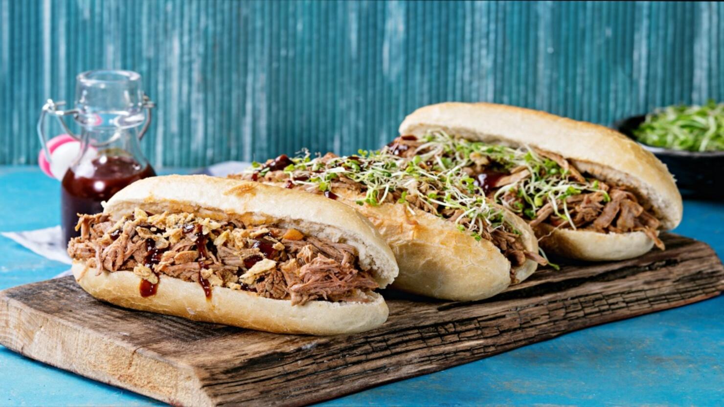 Variety of pulled pork sandwiches with meat, fried onion, green sprouts and bbq ketchup, served on wood cutting board with small bottle of tomato sauce over bright blue wooden background.