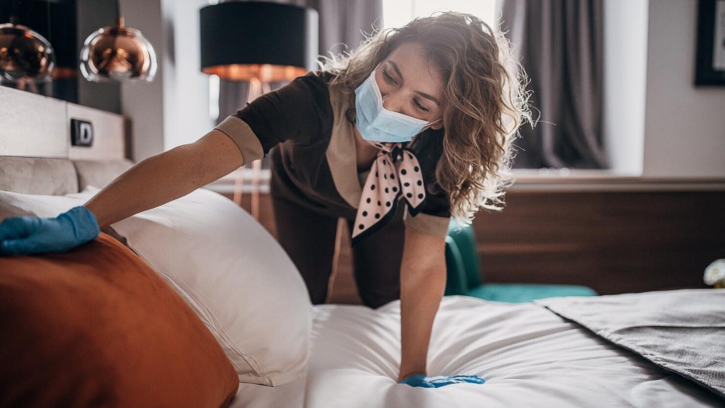 Beautiful maid with medical mask changing bed linen on the bed in a hotel room