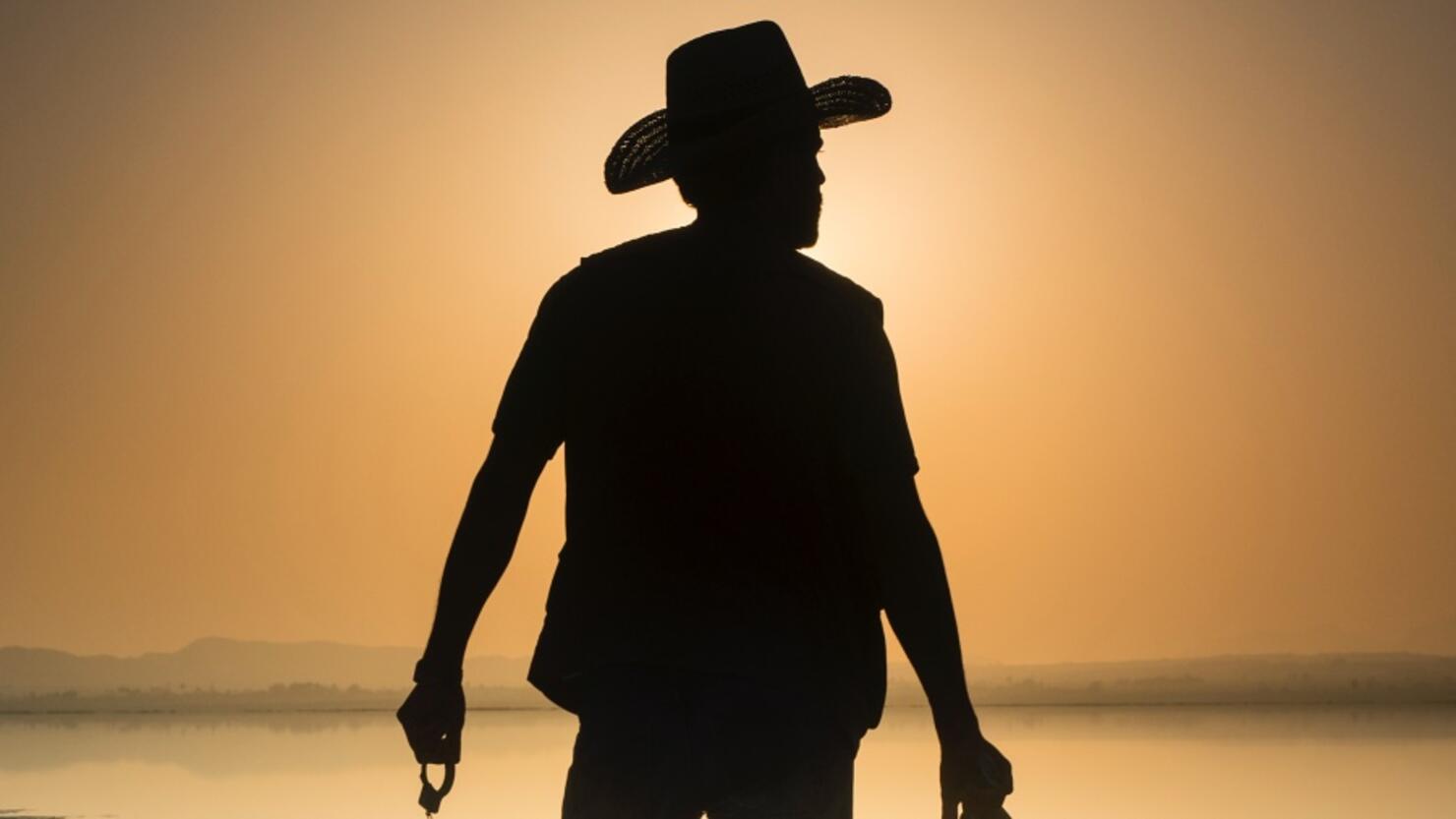 Silhouette of a man with cowboy clothes and pistoala front of a lake