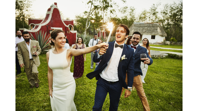 Bride and groom holding sparkler while celebrating during outdoor wedding reception with friends