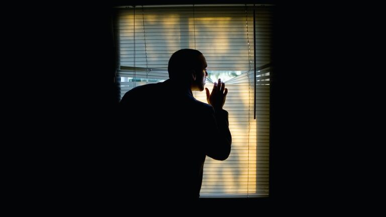 A man peeking outdoors through the blinds of a darkened room