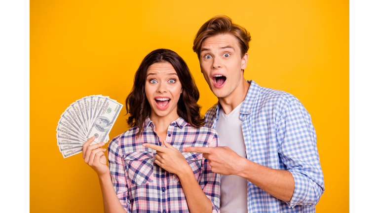 Close up photo of pair pack of bucks in hands he him his she her lady boy mouth opened luck shouting showing fingers on money wearing casual plaid shirts outfit isolated on yellow orange background
