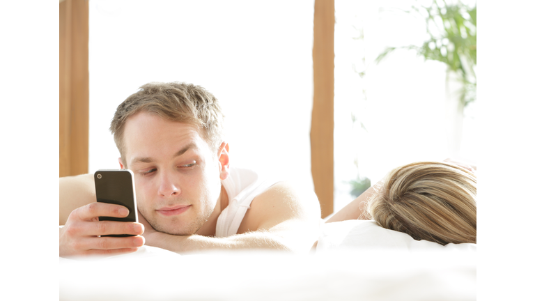 Man with phone in bed, looking at woman asleep