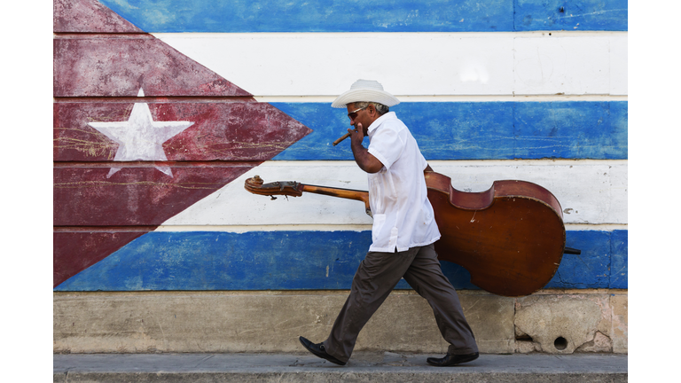 Hispanic musician carrying upright bass in front of Cuban flag mural