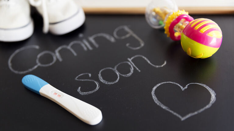 Waiting for labour, maternity concept. "Coming soon" words on black chalkboard with positive pregnancy test, white baby shoes and rattle