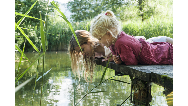 Mother and daughter lying on jetty at a lake looking at water