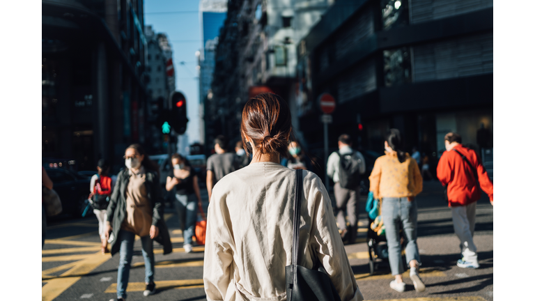 Rear view of young Asian woman commuting to work in city in the fresh morning, crossing street amidst crowd of pedestrians. Daily life and routine of a businesswoman