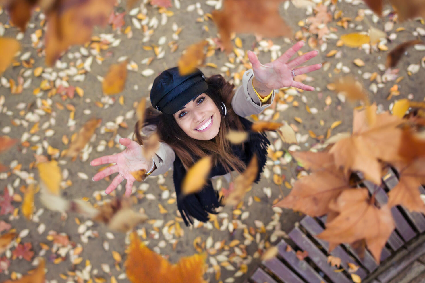 Pretty young woman looking to the sky with arms raised as leaves fall from the trees in the park in autumn.