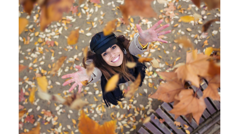 Pretty young woman looking to the sky with arms raised as leaves fall from the trees in the park in autumn.