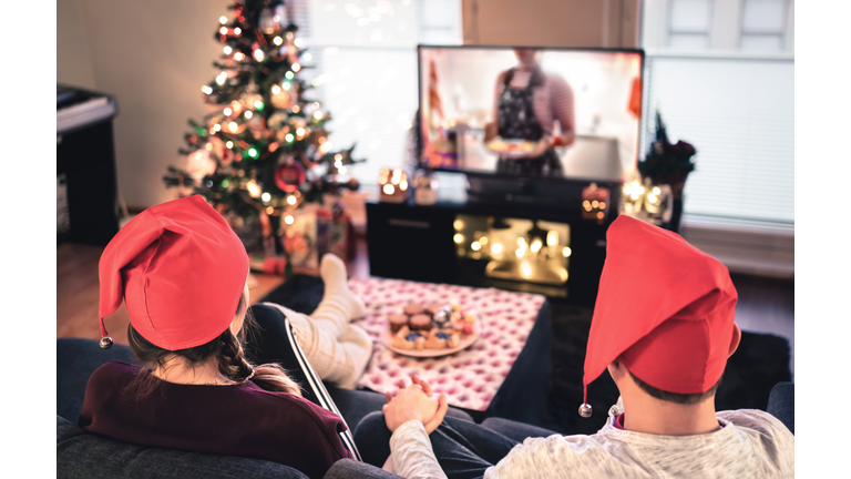 Couple watching tv on Christmas. Happy family holiday at home. Man and woman on couch relaxing with tree, decorations, lights, candles and television. Modern city celebrations.