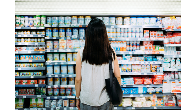 Rear view of young Asian mother groceries shopping for baby products in a supermarket. She is standing in front of the baby product aisle and have no idea which product to choose from