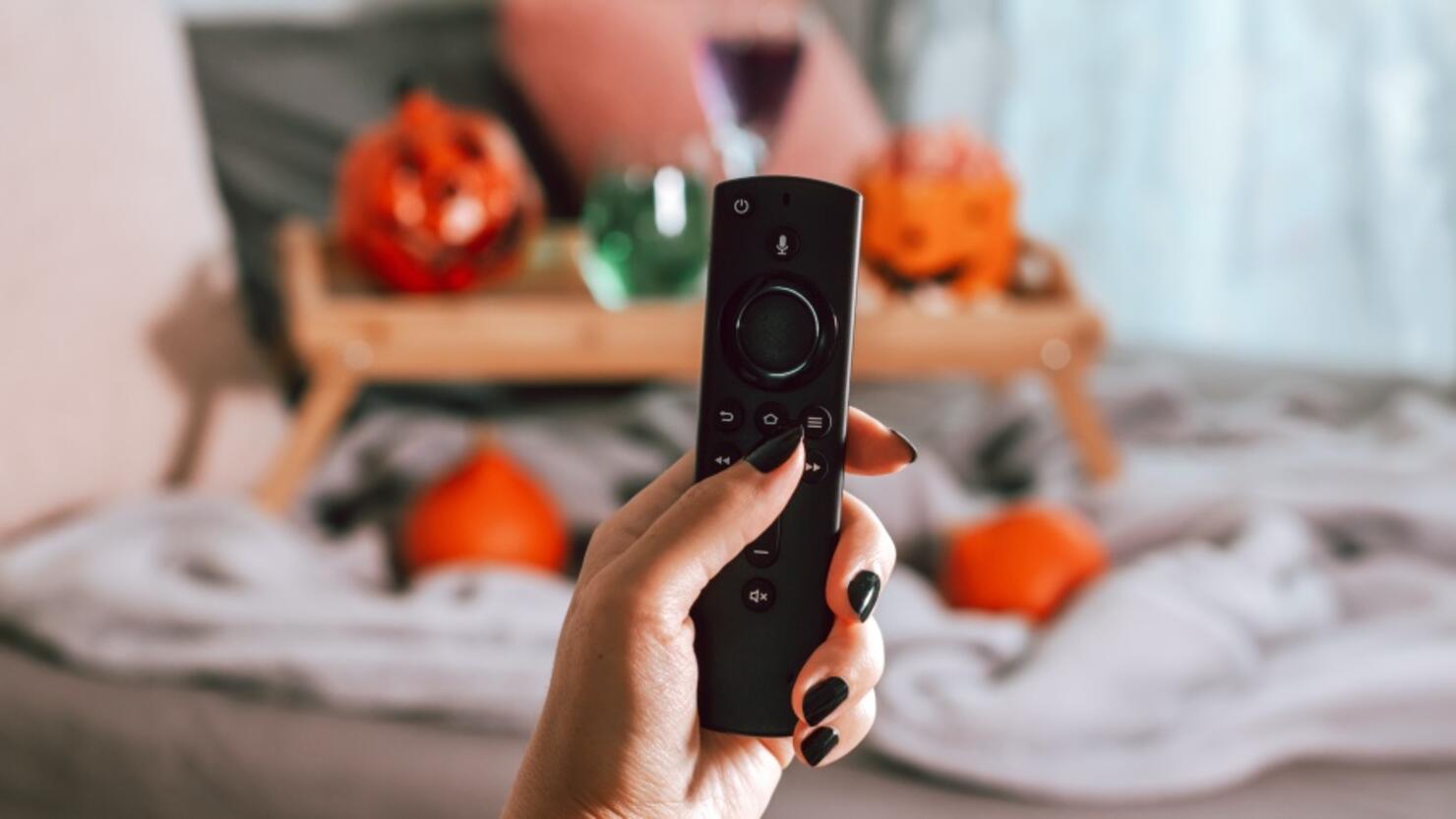 Woman hand with black manicure holding tv remote with Halloween decor in background, Halloween movie night concept