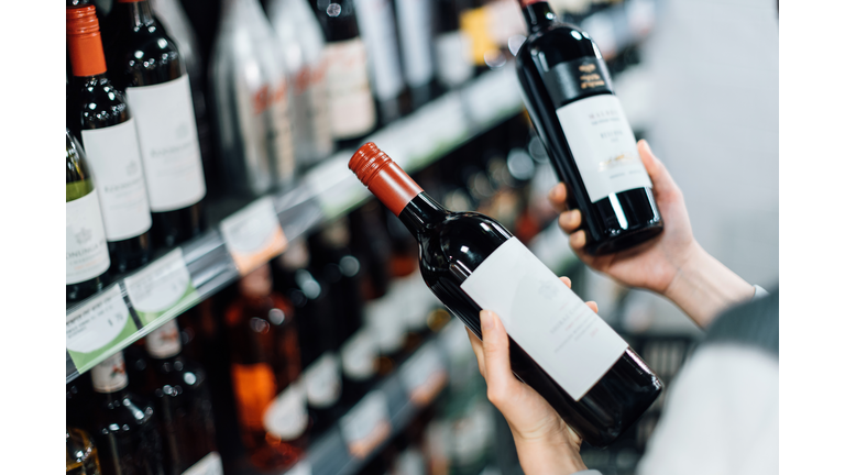Over the shoulder view of woman walking through liquor aisle and choosing bottles of red wine from the shelf in a supermarket