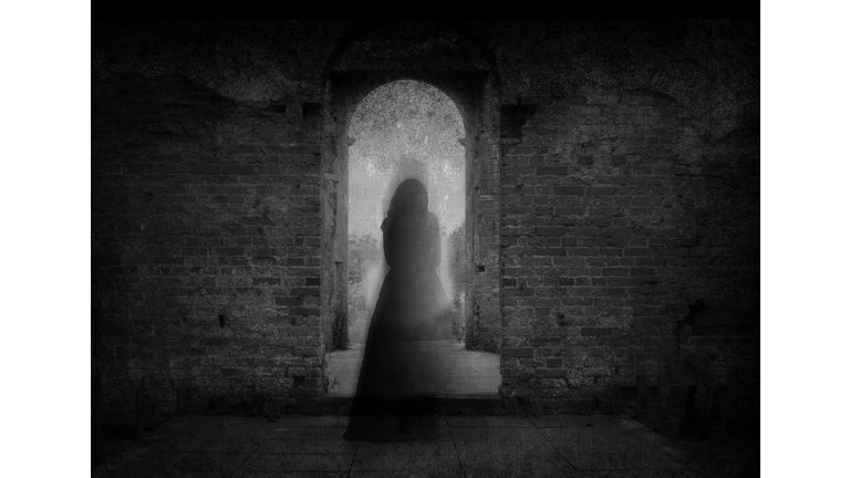 A spooky ghost of a woman in a dress, back to camera, framed by the archway of an old building. With a grunge, vintage, blurred edit.