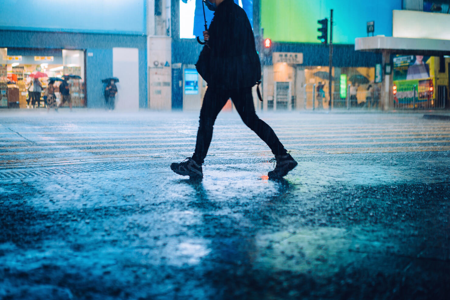 Silhouette of a man carrying umbrella walking in downtown city street against the reflection of glowing neon lights and city buildings in heavy rain at night
